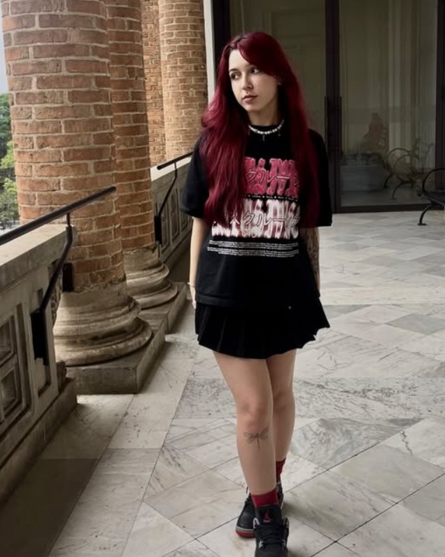 Luize entre colunas de um casarão, camiseta preta e saia